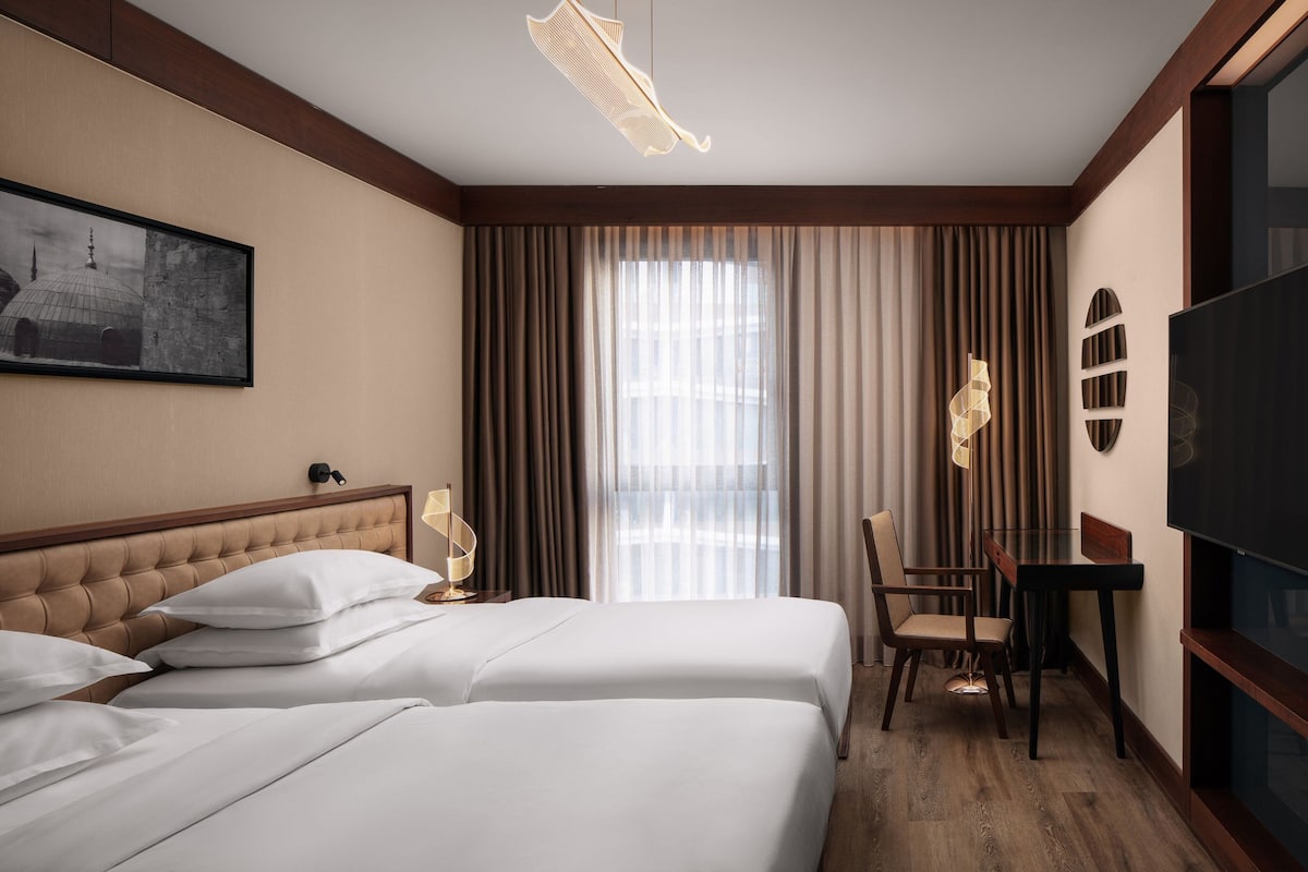 Habitación de hotel con dos camas, un escritorio, una silla, un televisor y un ventanal grande con cortinas.