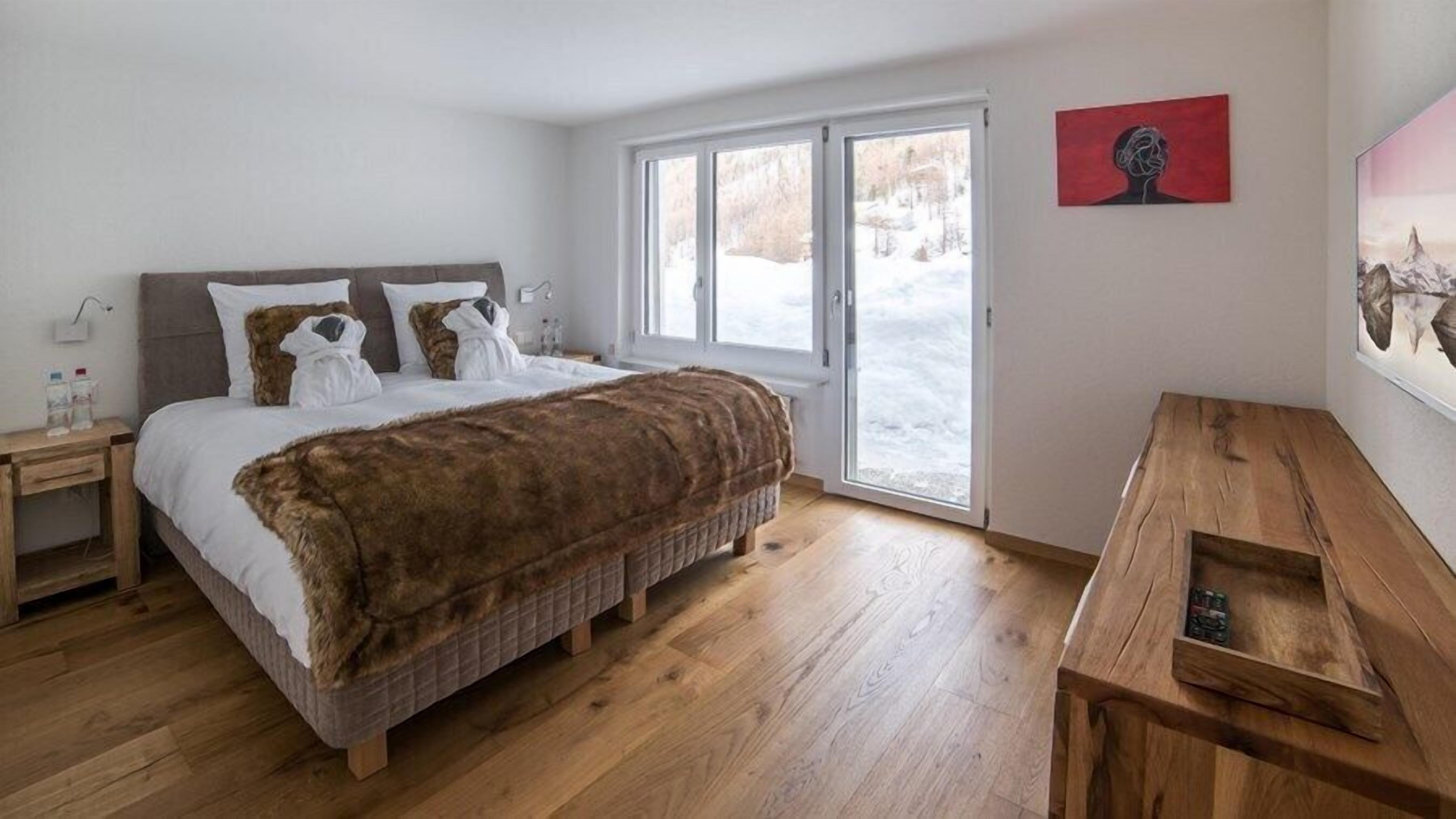 A bedroom with a bed, wooden floor, a bench, a window, and a painting on the wall.