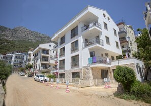 Un edificio de varios pisos con balcones, un camino de tierra en primer plano y montañas al fondo.