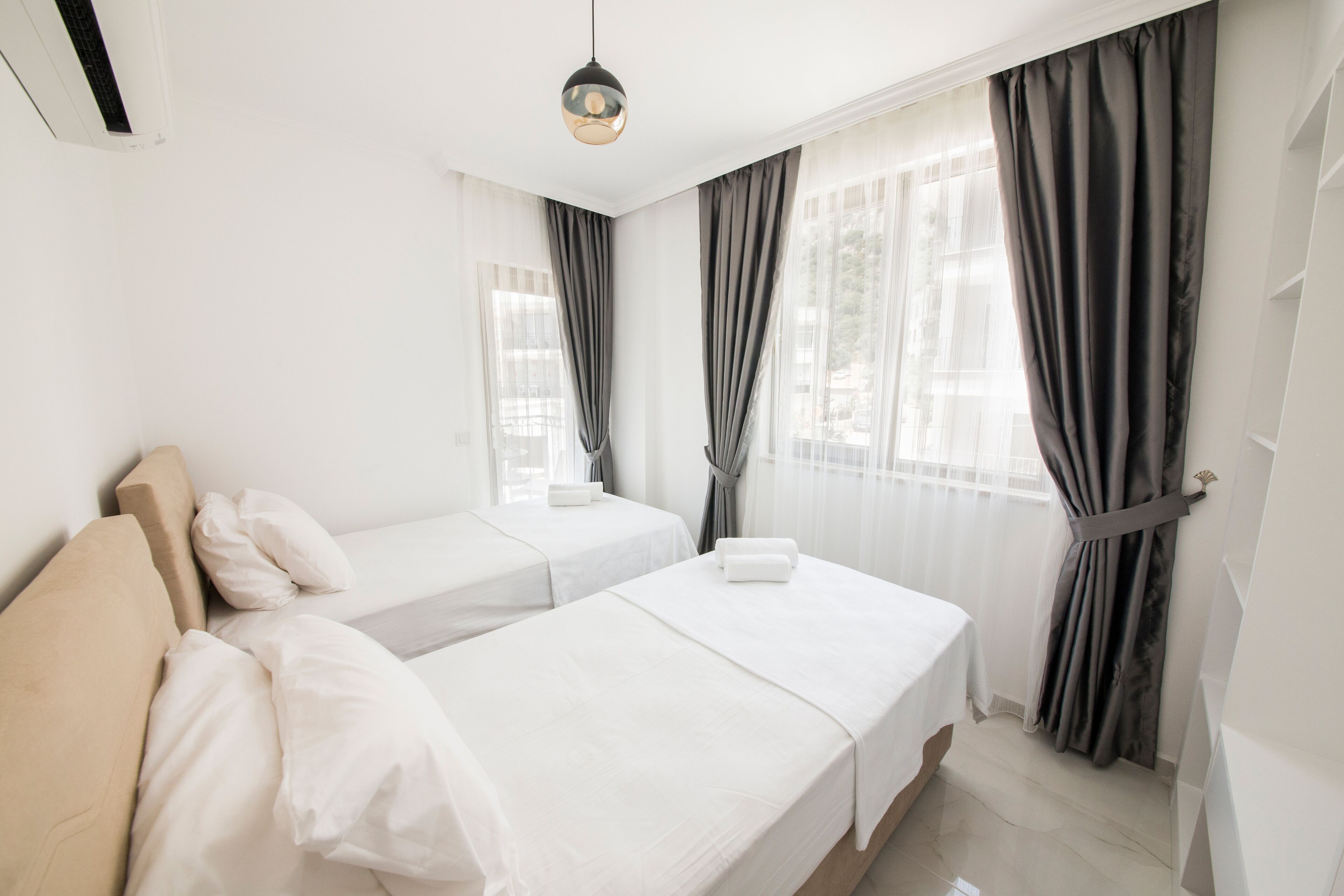A hotel room with two beds, a large window with curtains, a ceiling light, and a white wall.