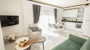 A modern living room with a green sofa, a white armchair, and a dining area with a table and chairs. A kitchen with white cabinets and appliances is visible in the background. - Cedrus Suites (Kas)