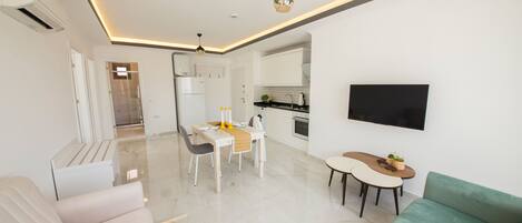 A modern living room with a sofa, a dining area with a table and chairs, a kitchen with a TV mounted on the wall, and a hallway leading to another room.