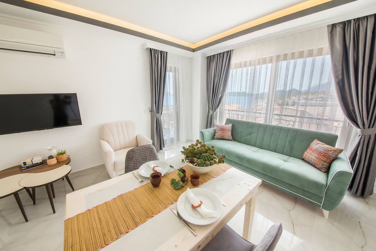 A modern living room with a dining table set for two, a green sofa, and a flat-screen TV mounted on the wall.