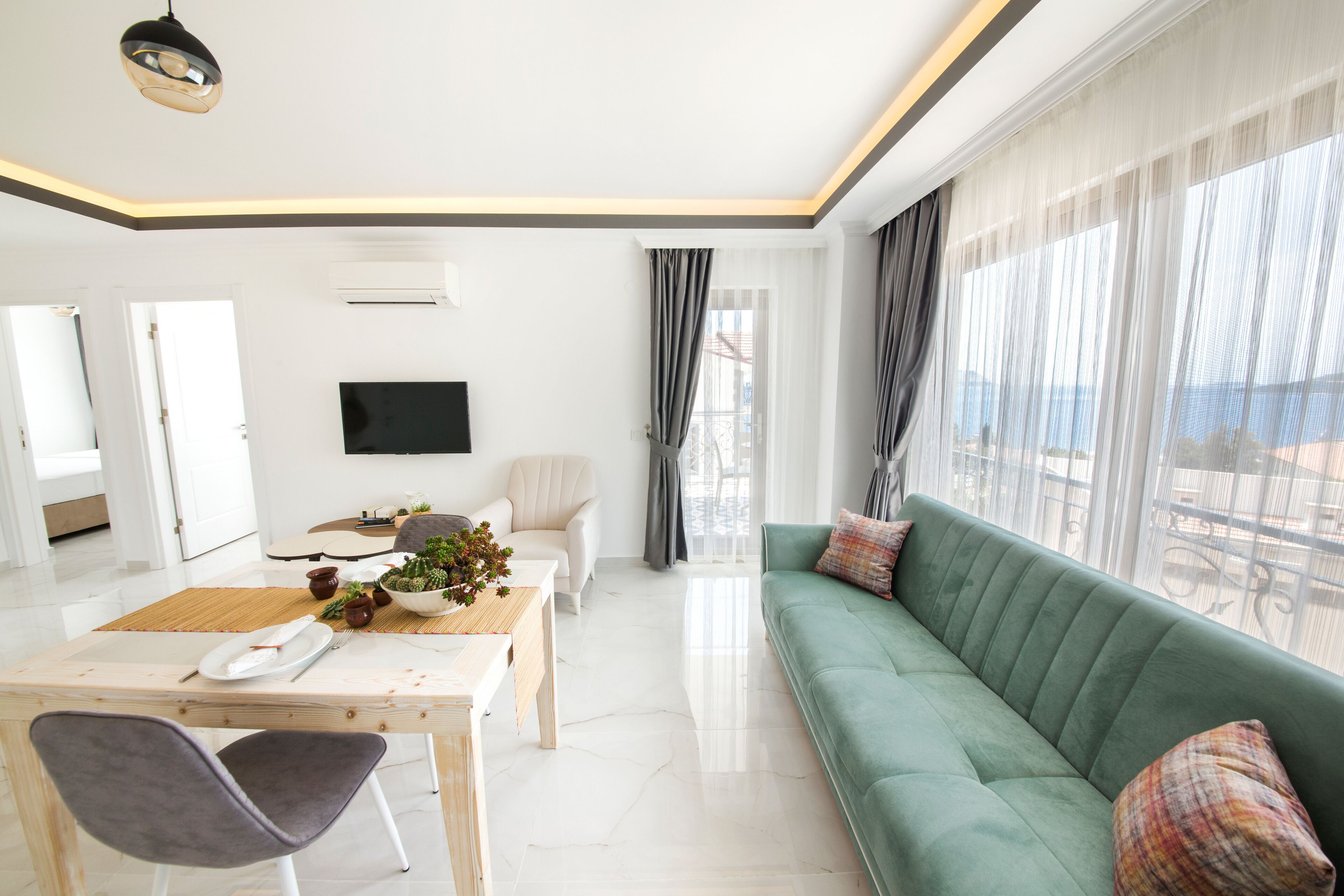 Ein modernes Wohnzimmer mit einem grünen Sofa, einem Esstisch mit Stühlen, einem Flachbildfernseher und einem großen Fenster mit Vorhängen.