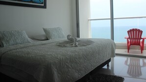 A bedroom with a large bed, a red chair, and a view of the sea through a large window. - Palmetto Eliptic 1002 (Cartagena)