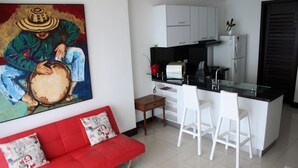 A modern kitchen with white cabinets, a black countertop, and a refrigerator. There is a red sofa with patterned cushions and a small table with a remote control. - Palmetto Eliptic 1002 (Cartagena)