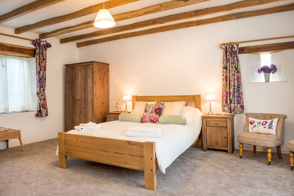 A bedroom with a wooden bed, two bedside tables with lamps, a wooden wardrobe, a chair, and a window with curtains.