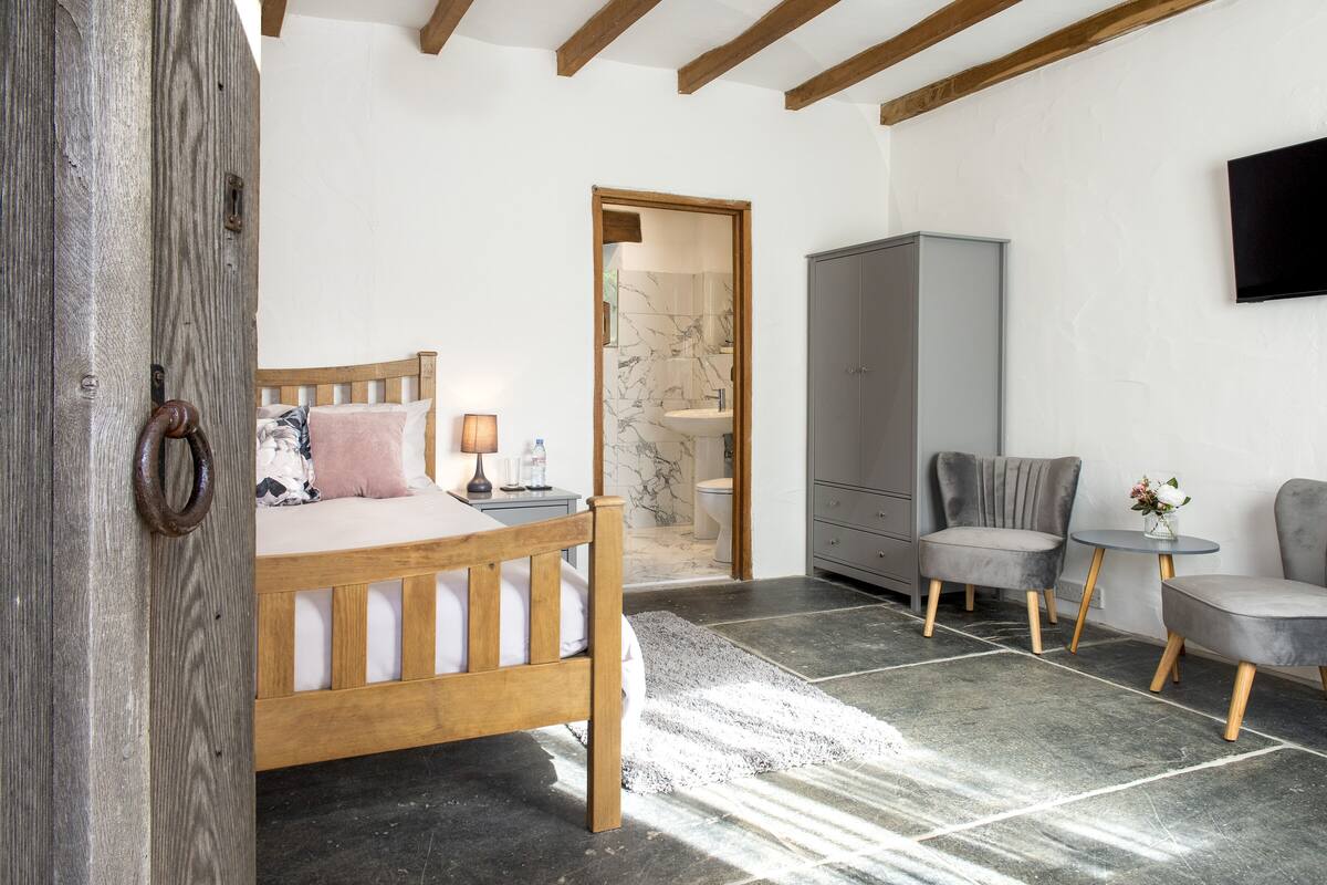 A bedroom with a wooden bed, a grey wardrobe, a grey armchair, a round table, and a TV mounted on the wall.