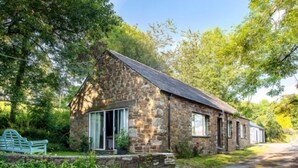 A stone cottage with a sloped roof, large windows, and a bench in the garden. - Coombe Farm (Looe)