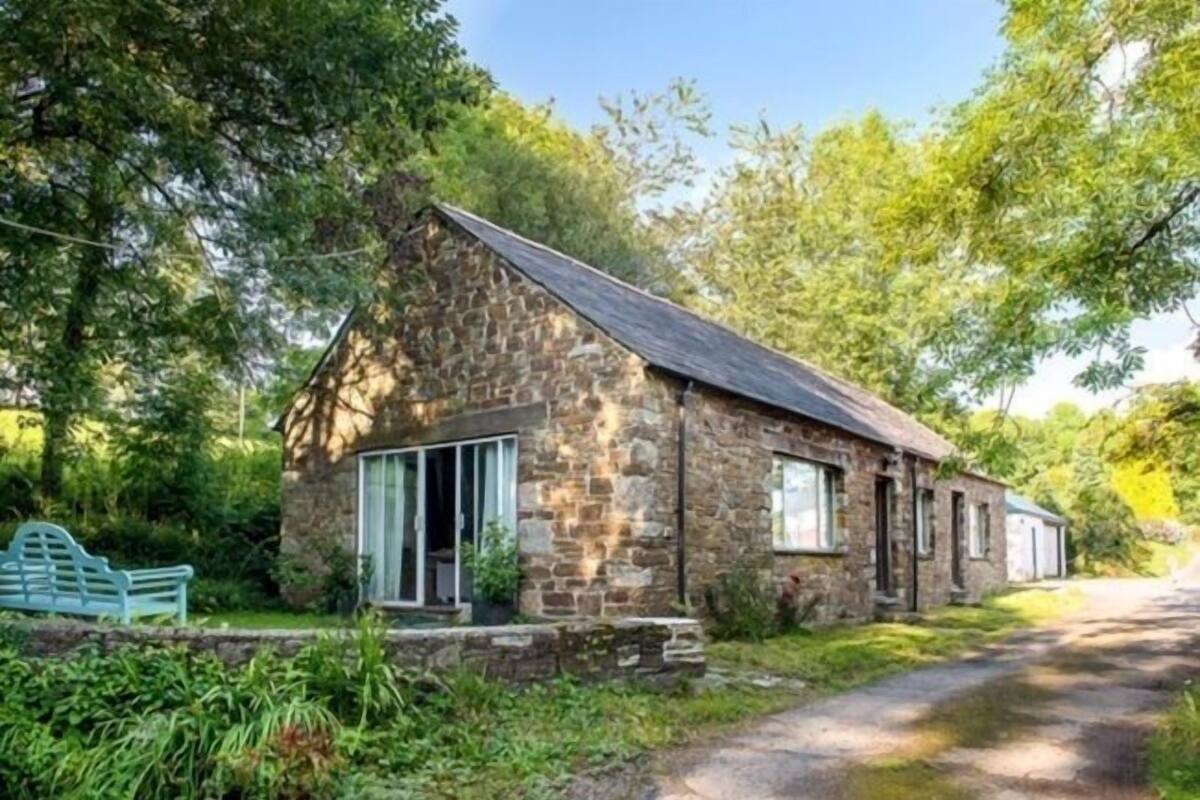 A stone cottage with a sloped roof, large windows, and a bench in the garden.