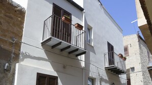 Un edificio de dos pisos con fachada blanca, un balcón pequeño con barandilla metálica y una puerta con un ventanal.