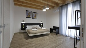 Un dormitorio con piso de madera, una cama con ropa de cama blanca, dos mesitas de noche, un escritorio y una ventana con cortinas.