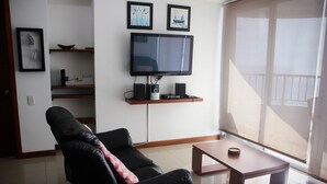 Een woonkamer met een zwarte leren bank, een salontafel, een flatscreen televisie en planken met decoratieve objecten.