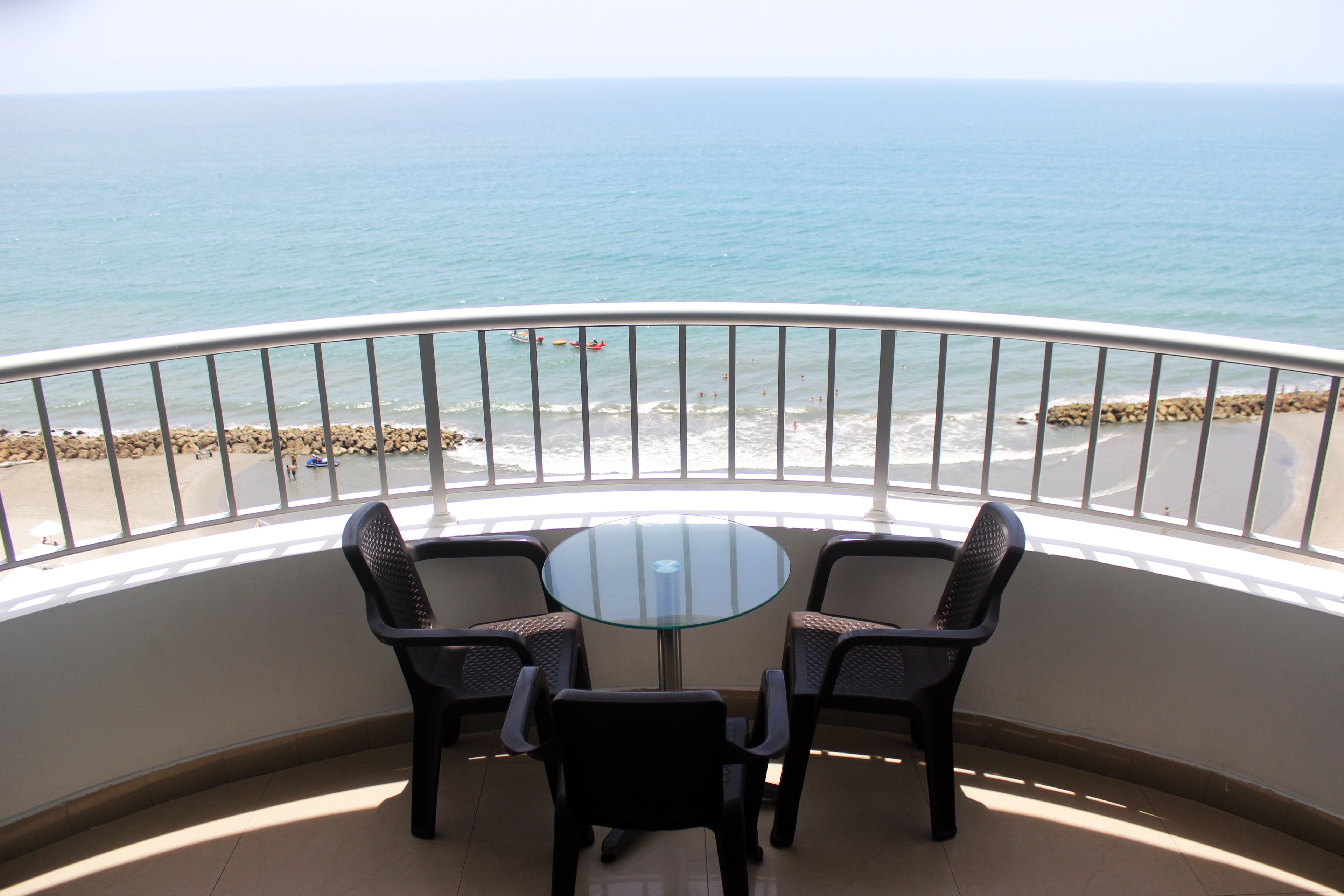 Ein Balkon mit Glastisch und vier Stühlen, mit Blick auf einen Strand mit felsigem Ufer und ruhiger See.