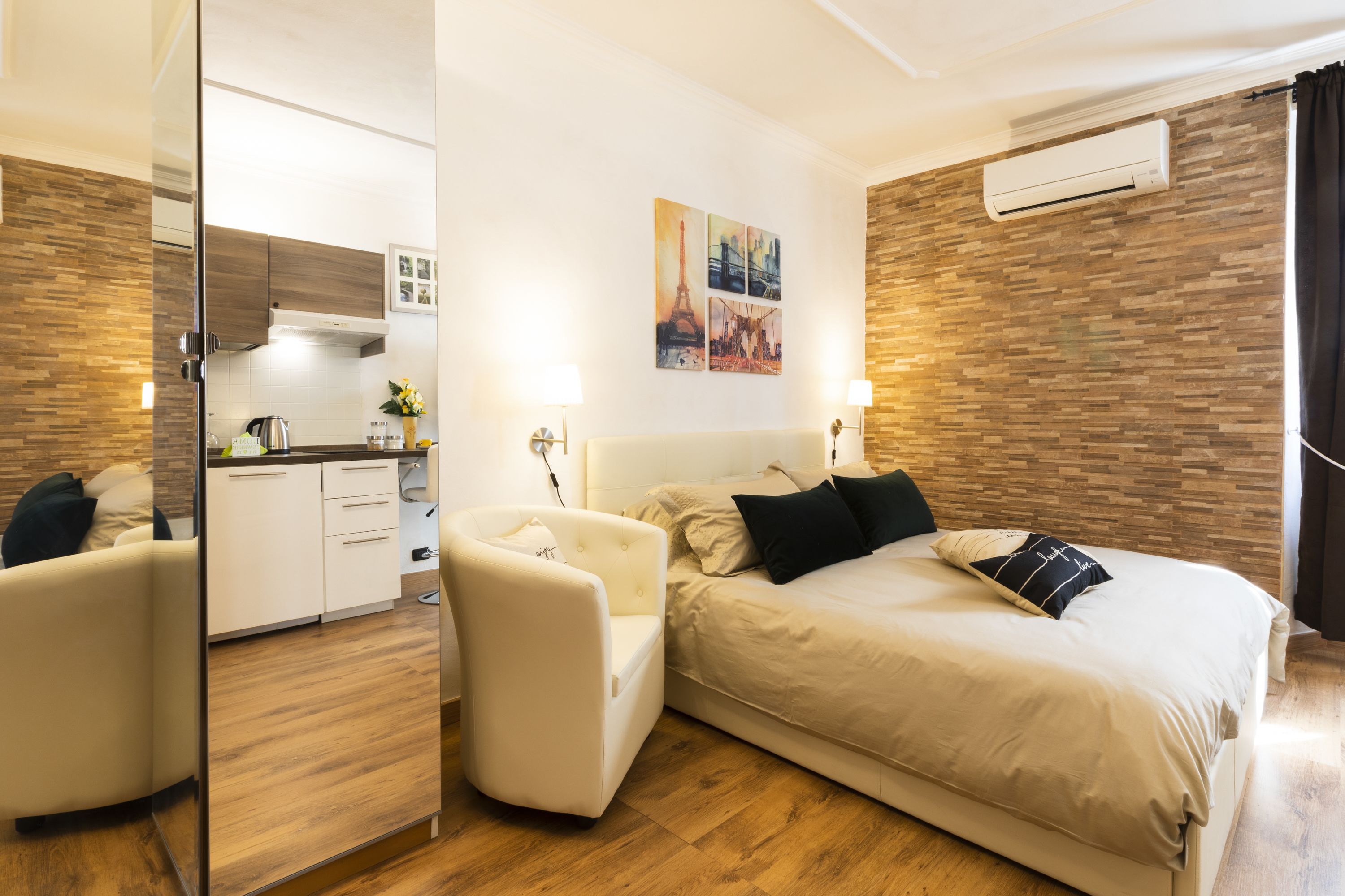 A modern hotel room with a bed, a chair, a kitchen area, and a mirror.