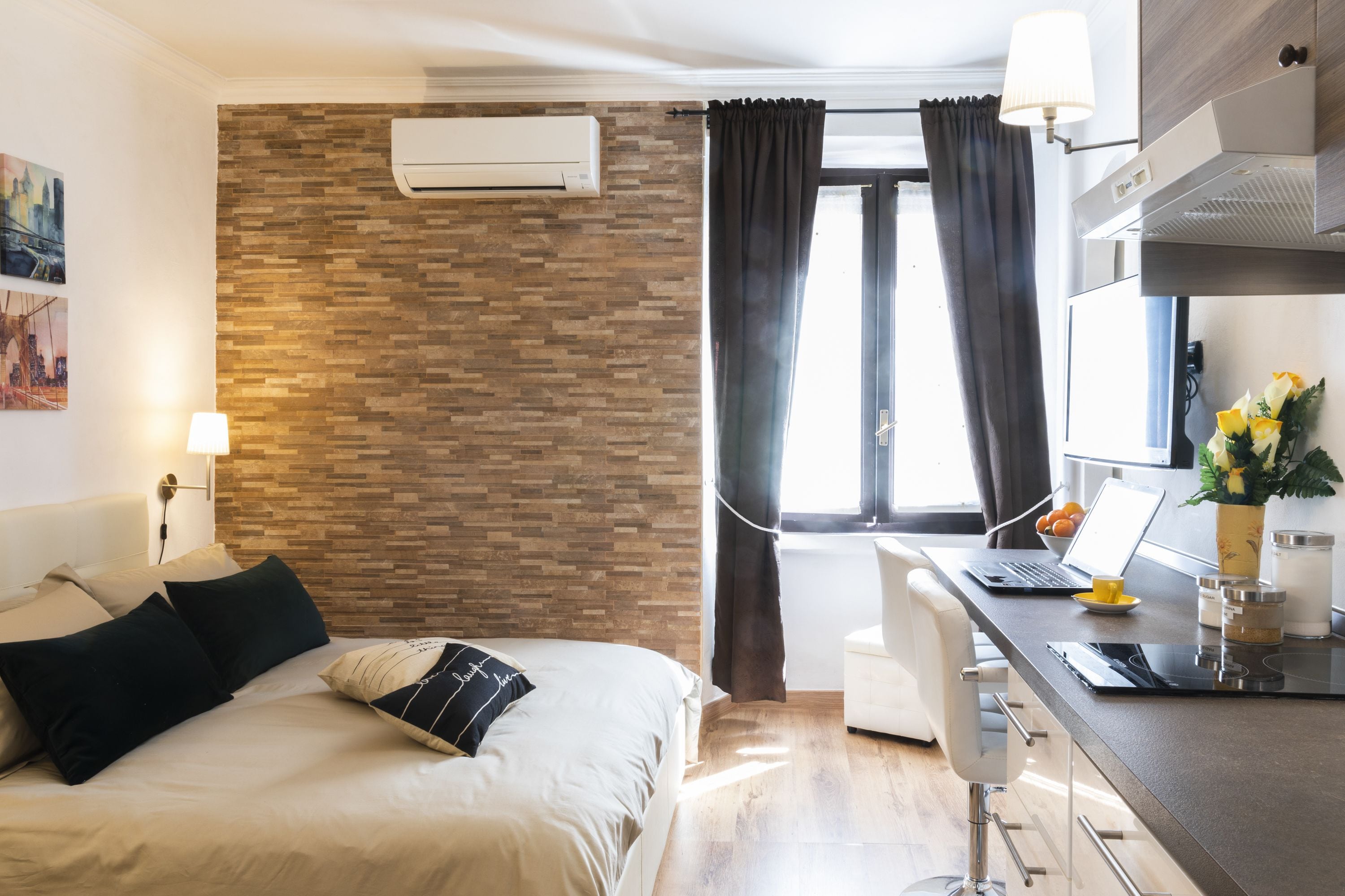 Een moderne hotelkamer met een accentmuur in baksteedesign, een bed met donkere en lichte kussens, een bureau met een laptop, een vaas met bloemen en een raam met gordijnen.