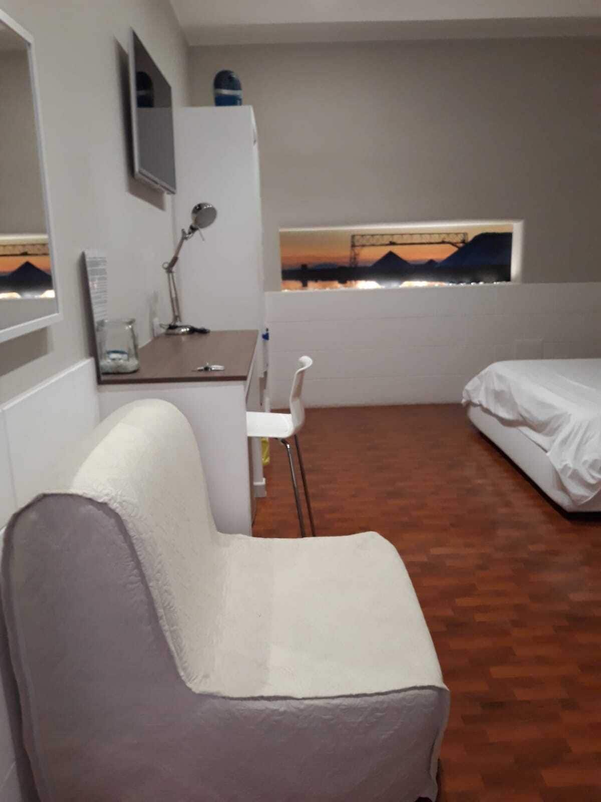 Een kleine hotelkamer met een bed, een bureau met stoel, een klein tafeltje, een spiegel en een raam met uitzicht op een zonsondergang.
