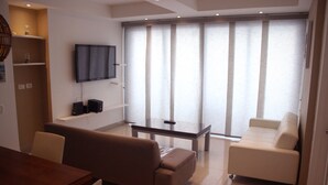 Un salon moderne avec un canapé marron, un fauteuil blanc, une table basse en verre et un téléviseur à écran plat fixé au mur.