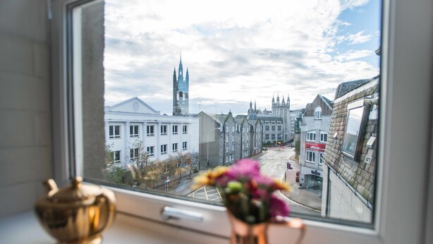 Une vue par la fenêtre sur un paysage urbain, avec des bâtiments historiques et le clocher d’une église.