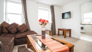 Un salon avec un canapé marron, une table basse en verre, une table d’appoint en bois, un téléviseur fixé au mur et un lave-linge à l’arrière-plan.