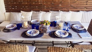 Una mesa de comedor dispuesta con platos azules y blancos, vasos y velas, rodeada de sillas de mimbre y un fondo de madera.