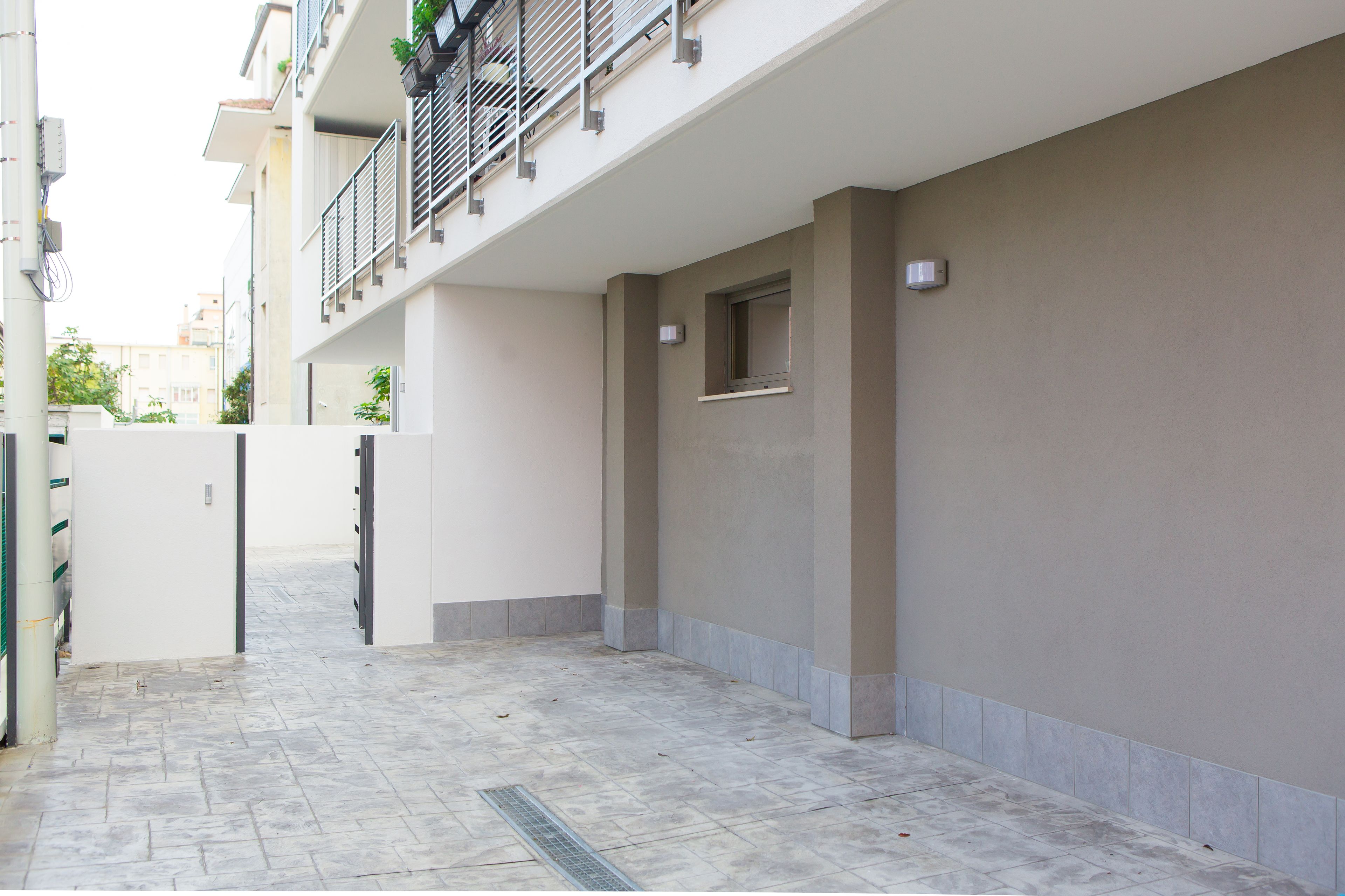 A clean, modern residential building with a tiled courtyard, a balcony with metal railings, and a small window on the right wall.