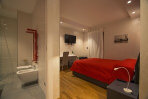 A modern bedroom with a red bedspread, a desk with a chair, a TV mounted on the wall, and a shower area. - Le Mie Città (Pescara)