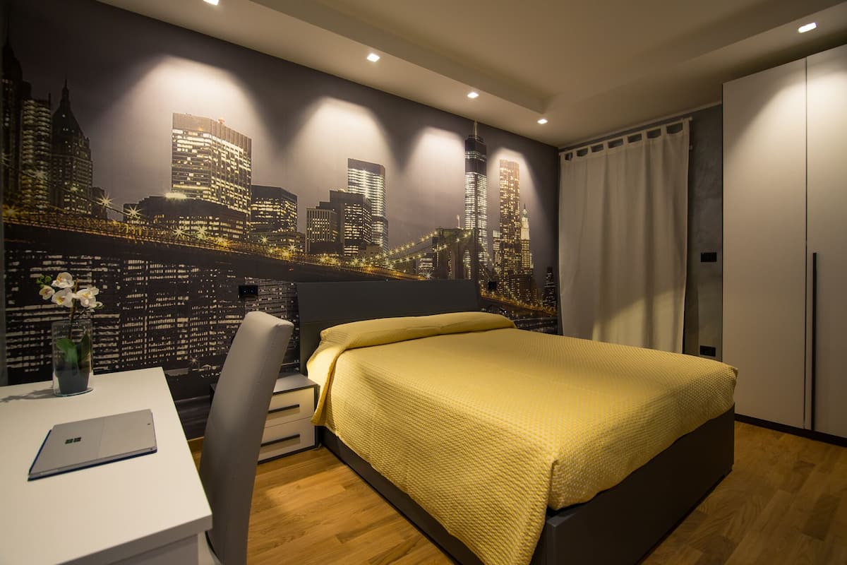 A modern bedroom with a cityscape mural, a bed with a yellow blanket, a desk with a laptop, and a chair.