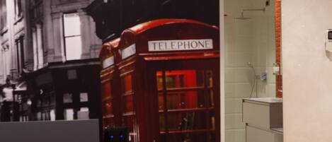 A red telephone booth with a sign reading "TELEPHONE" in a modern bathroom with a shower and a sink.