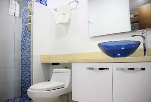 A bathroom with a white toilet, a blue mosaic shower wall, a white sink with a blue bowl, and a white cabinet. - Palmetto 1902 (Cartagena)