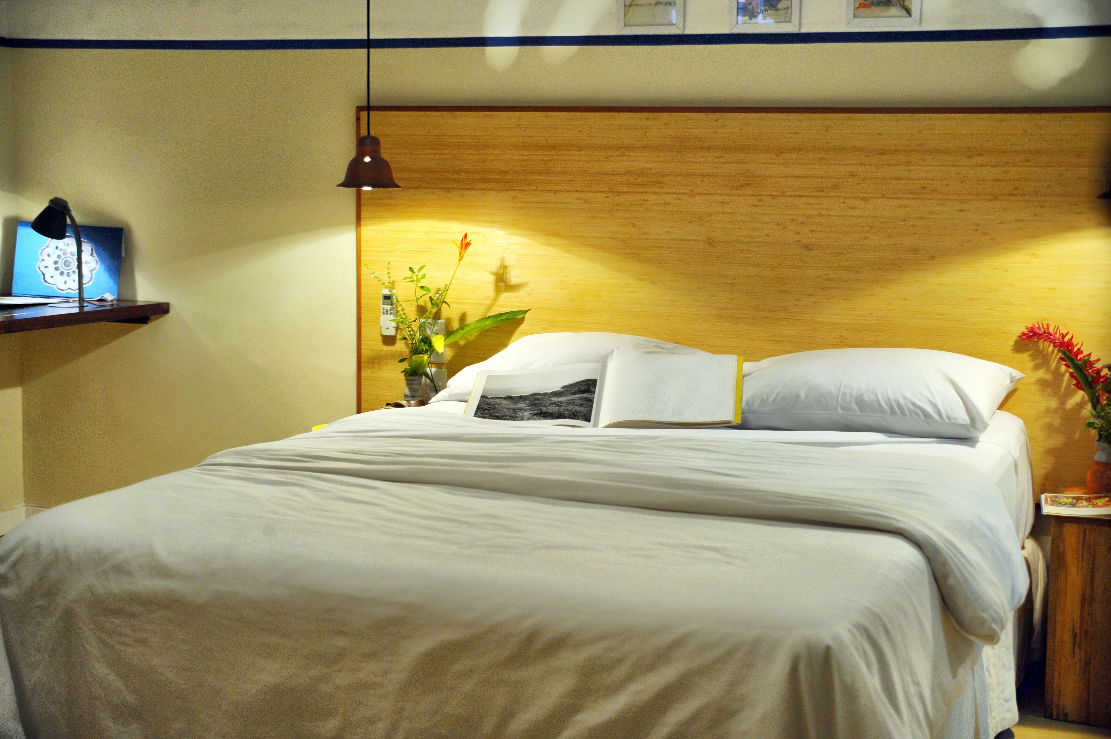 A neatly made bed with white linens, a bedside lamp, a small shelf with a book and a lamp, and a vase with flowers on the nightstand.