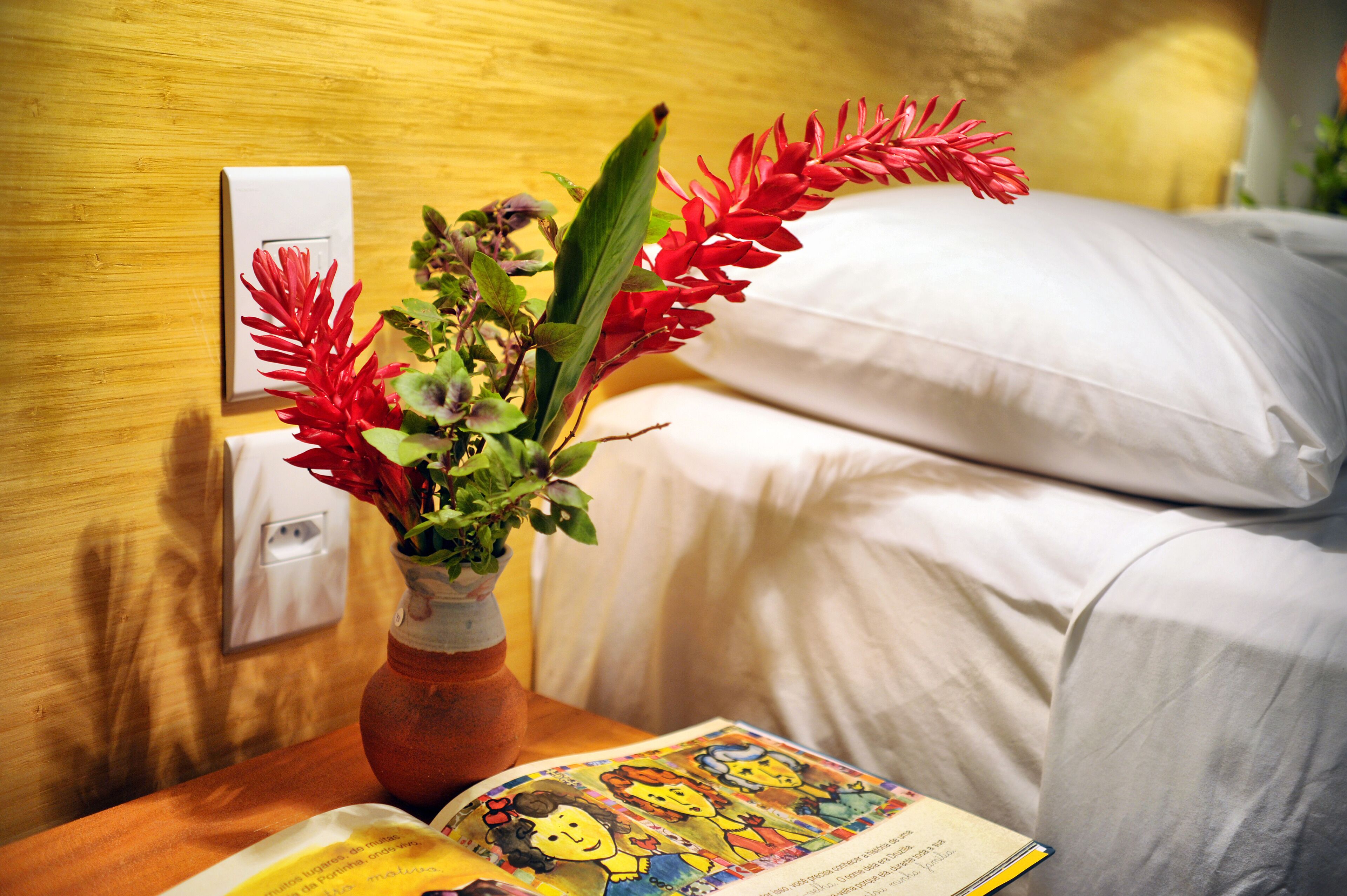 Ein Hotelzimmer mit einem hölzernen Kopfteil, einem weißen Kissen, einem Nachttisch mit einem Buch und einer Vase mit roten Blumen.