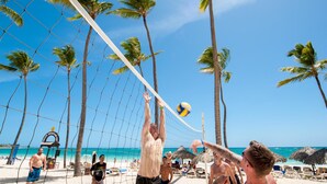 A beach volleyball game with players in shorts, palm trees, and a clear sky.