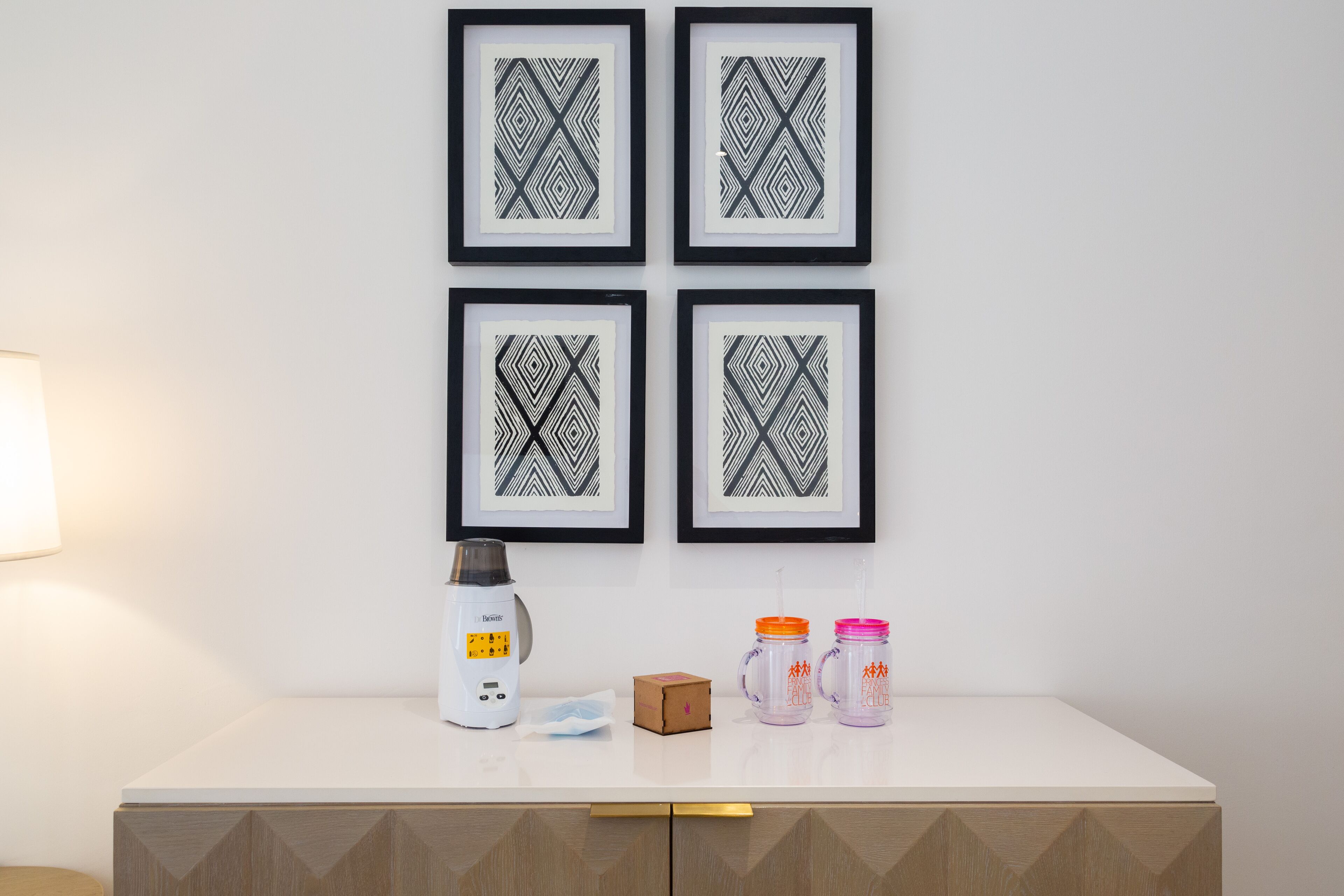 Un plan de travail blanc orné de quatre motifs géométriques encadrés, une machine à café blanche, deux récipients orange et rose, et une petite boîte marron.