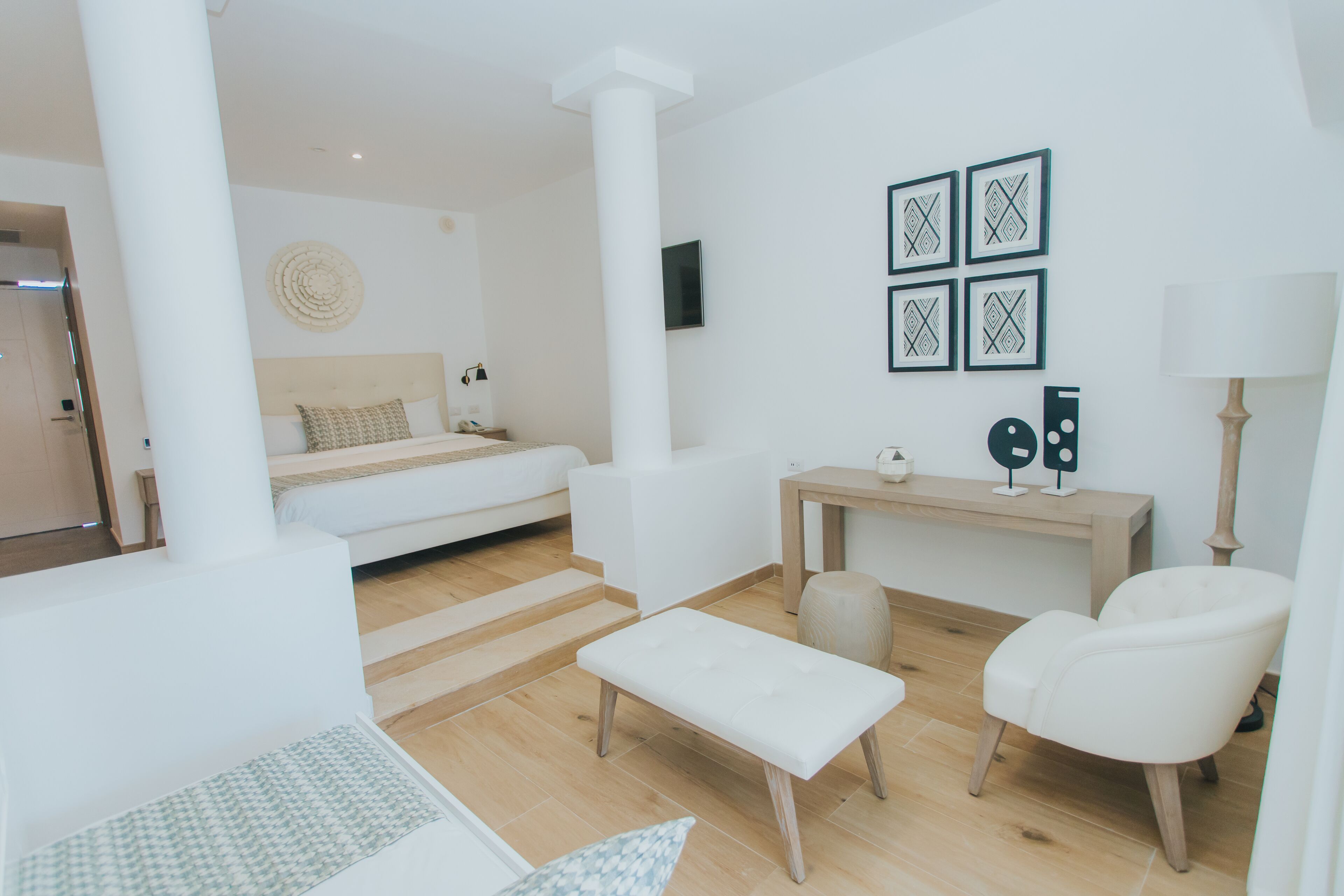 Una sala de estar moderna y minimalista con una cama, un banco, una silla, una mesita y un televisor fijado a la pared.