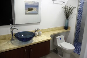 A bathroom with a blue glass bowl sink, a white toilet, a beige countertop, a vase with branches, and a painting of a boat on water. - Palmetto 2103 (Cartagena)