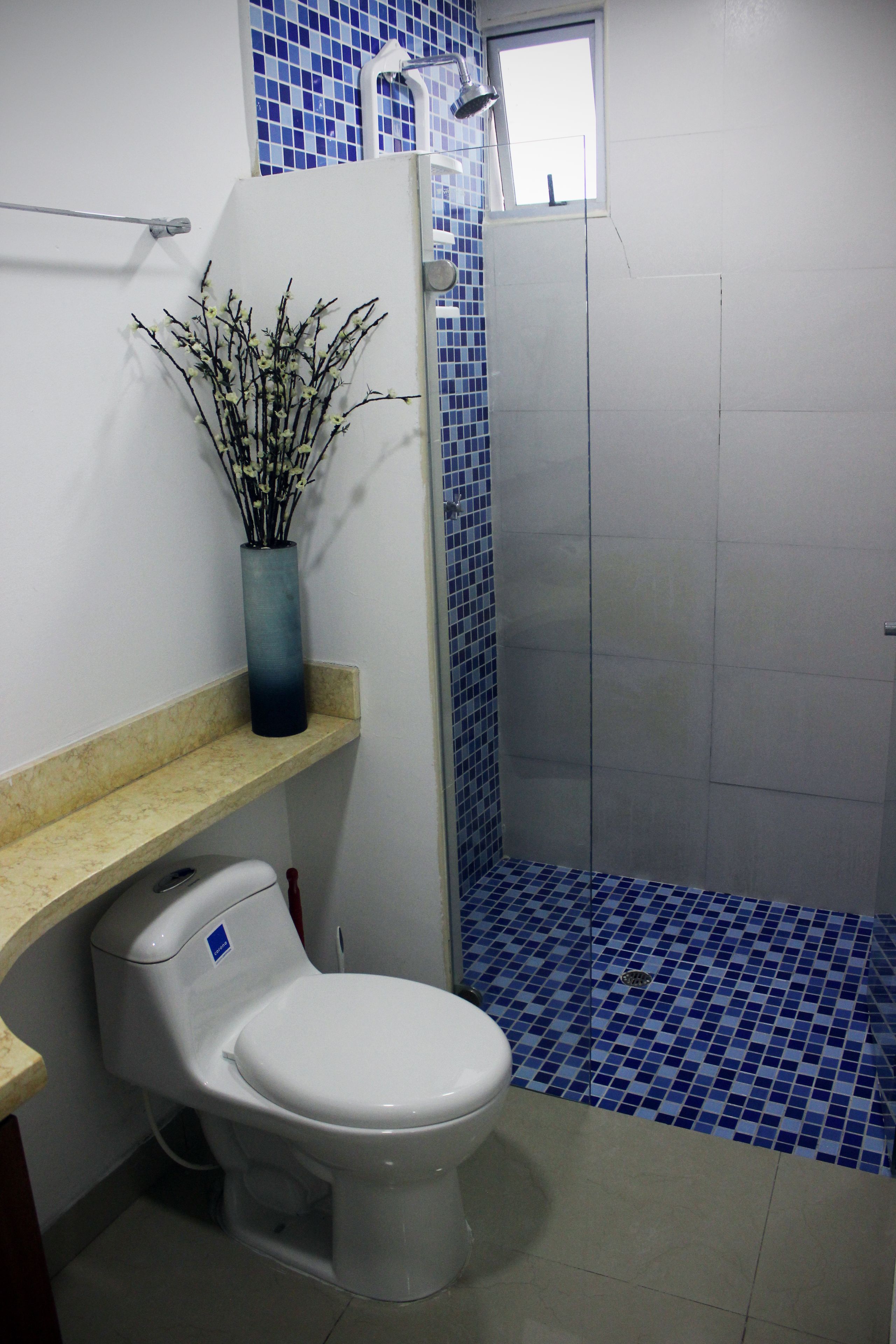 A bathroom with a white toilet, a blue-tiled shower area, and a vase with branches on a shelf.
