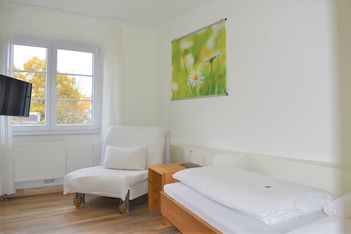 A hotel room with a single bed, a chair, a small table, a TV, and a window with curtains.
