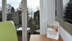 A room with a large window, a green chair, and a table with a book titled "Heute, hier, jetzt - Ein Reiseführer Durch Leben". - Hotel Lindenallee (Lindau)