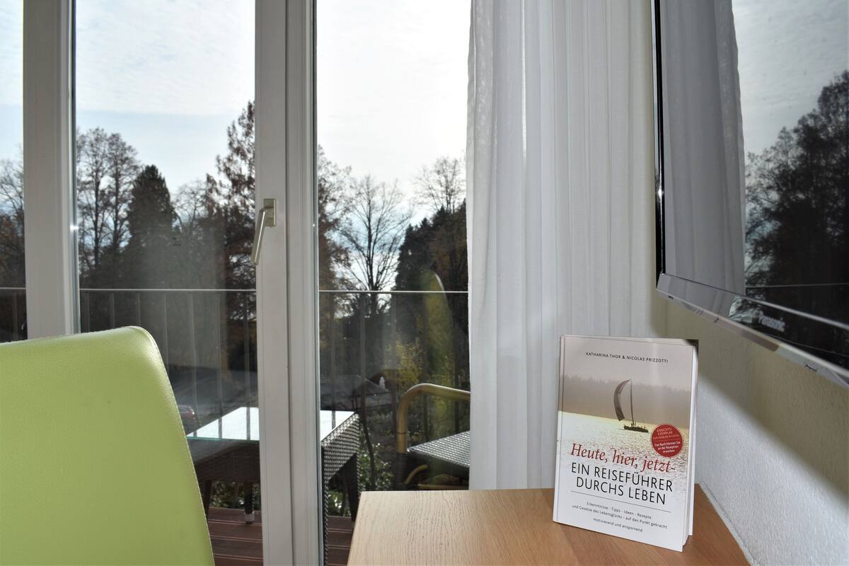 A room with a large window, a green chair, and a table with a book titled "Heute, hier, jetzt - Ein Reiseführer Durch Leben".