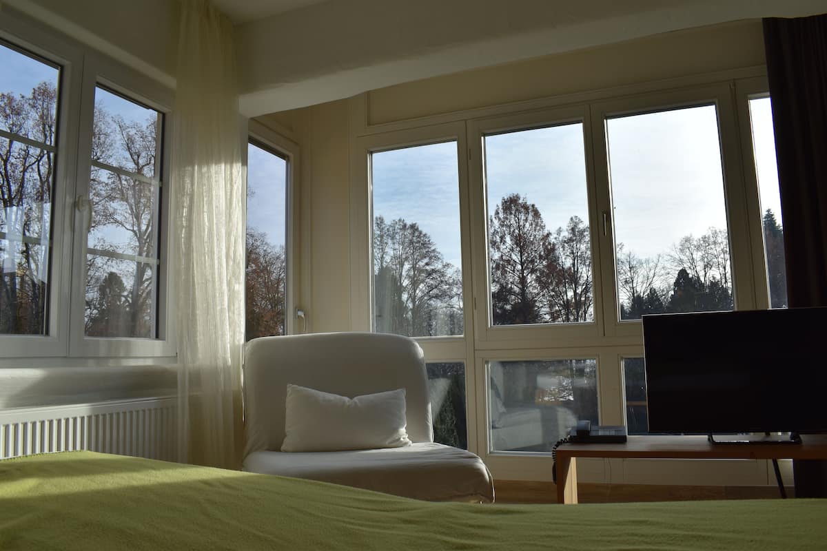 A hotel room with a bed, a chair, a desk with a computer, and large windows offering a view of trees and buildings.