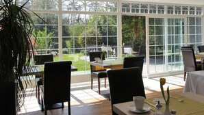 A dining area with large windows, tables, chairs, and a potted plant. - Hotel Lindenallee (Lindau)