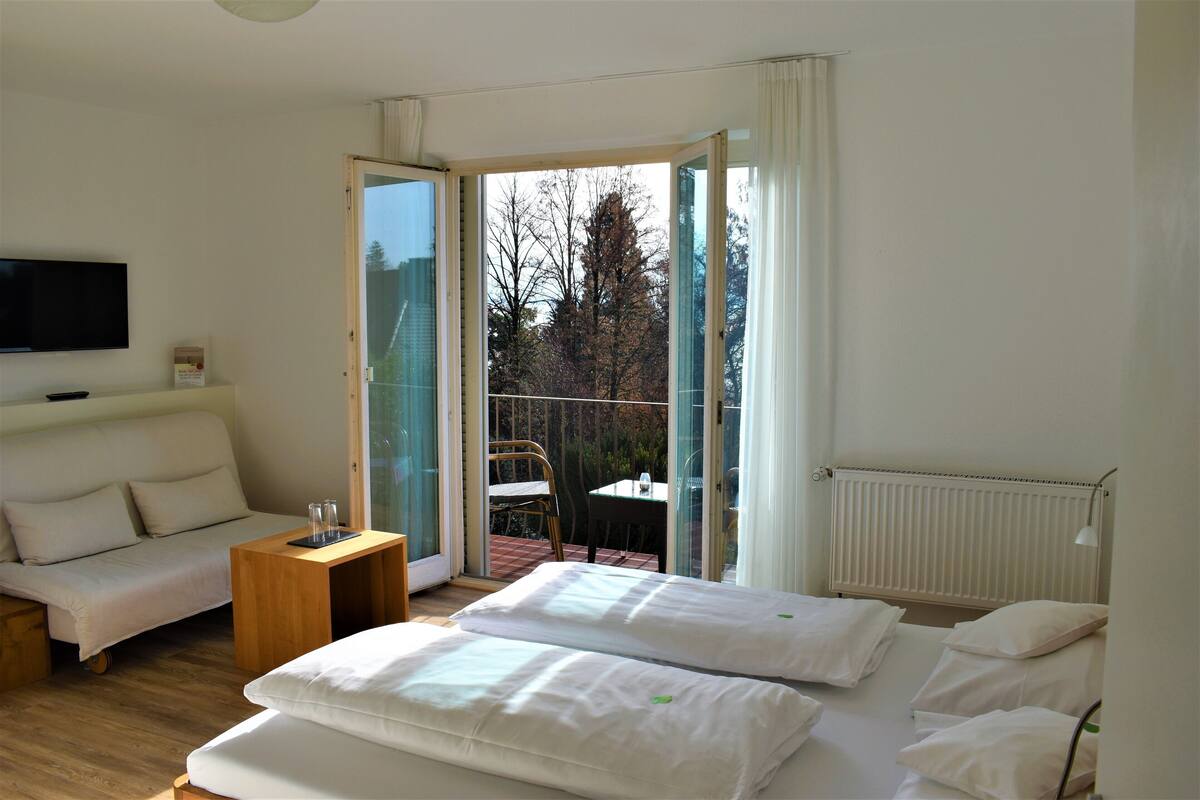 A hotel room with a bed, a sofa, a small table, a TV, and a sliding glass door leading to an outdoor area.