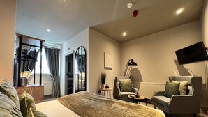 Una habitación de hotel moderna con una cama grande, un área de descanso con un sofá y sillones, un televisor de pantalla plana y una mesita de noche con lámpara.