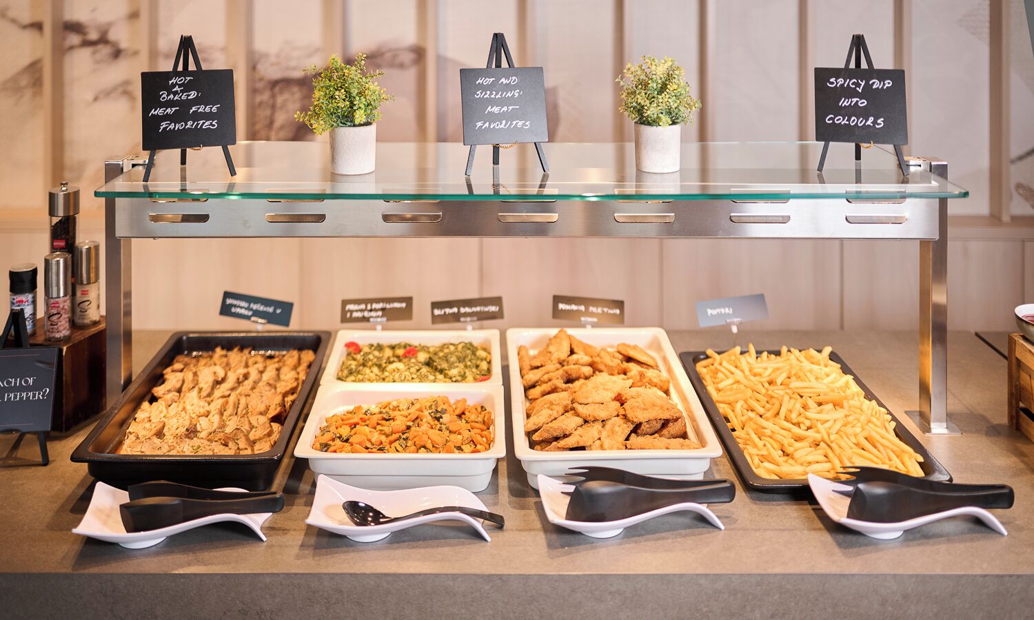 Un buffet avec des plateaux de nourriture clairement étiquetés, comprenant des options végétariennes, et un panneau indiquant une sauce épicée aux couleurs vives.