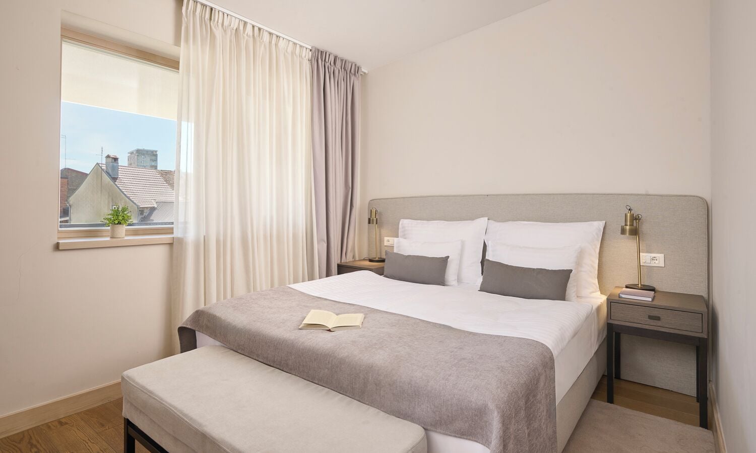 Un dormitorio con cama, mesitas de noche, una ventana con cortinas y vistas a edificios.