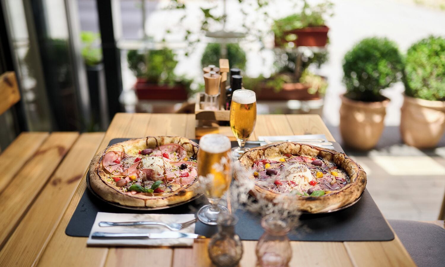 Two pizzas on a wooden table with a glass of beer and a pepper shaker.