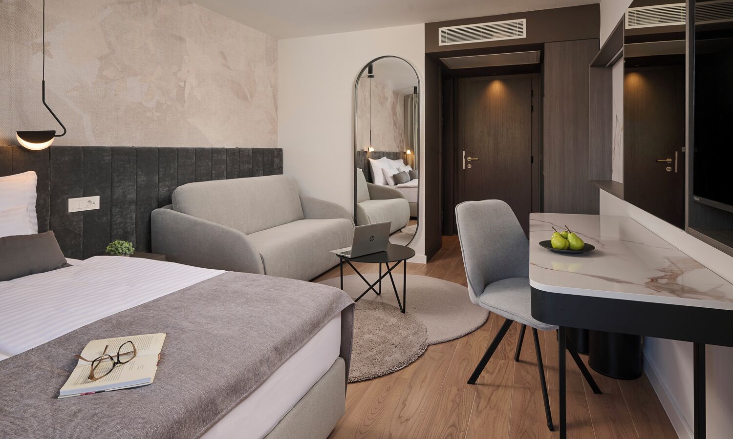 A modern hotel room with a bed, a sofa, a desk with a laptop, and a mirror.