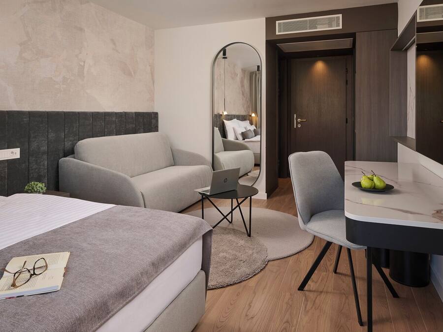 A modern hotel room with a bed, a sofa, a desk with a laptop, and a mirror.