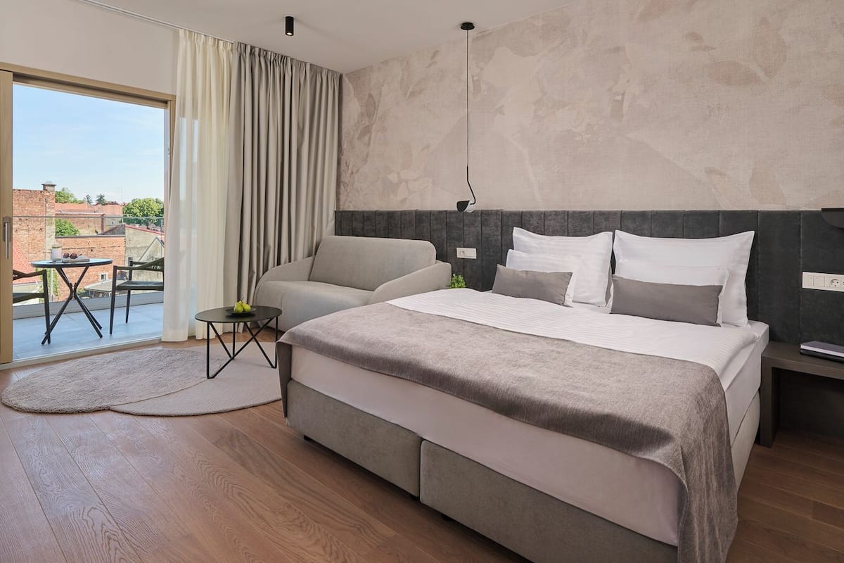 Quarto moderno com uma cama grande, um sofá, uma mesinha com frutas e vista para prédios através de uma porta de vidro.
